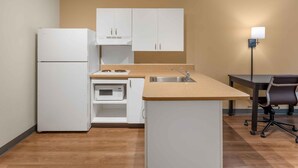 A compact kitchen with white cabinets, a microwave, a sink, and a dining area with a chair.