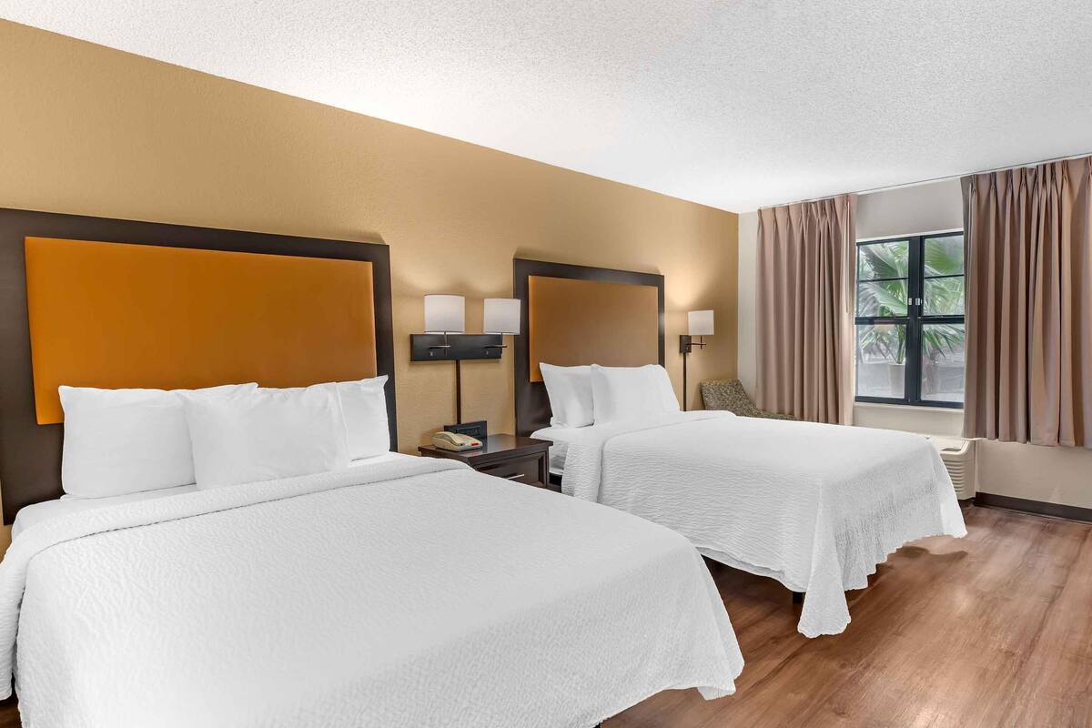 A hotel room with two beds, a large window with curtains, and a wall-mounted lamp.