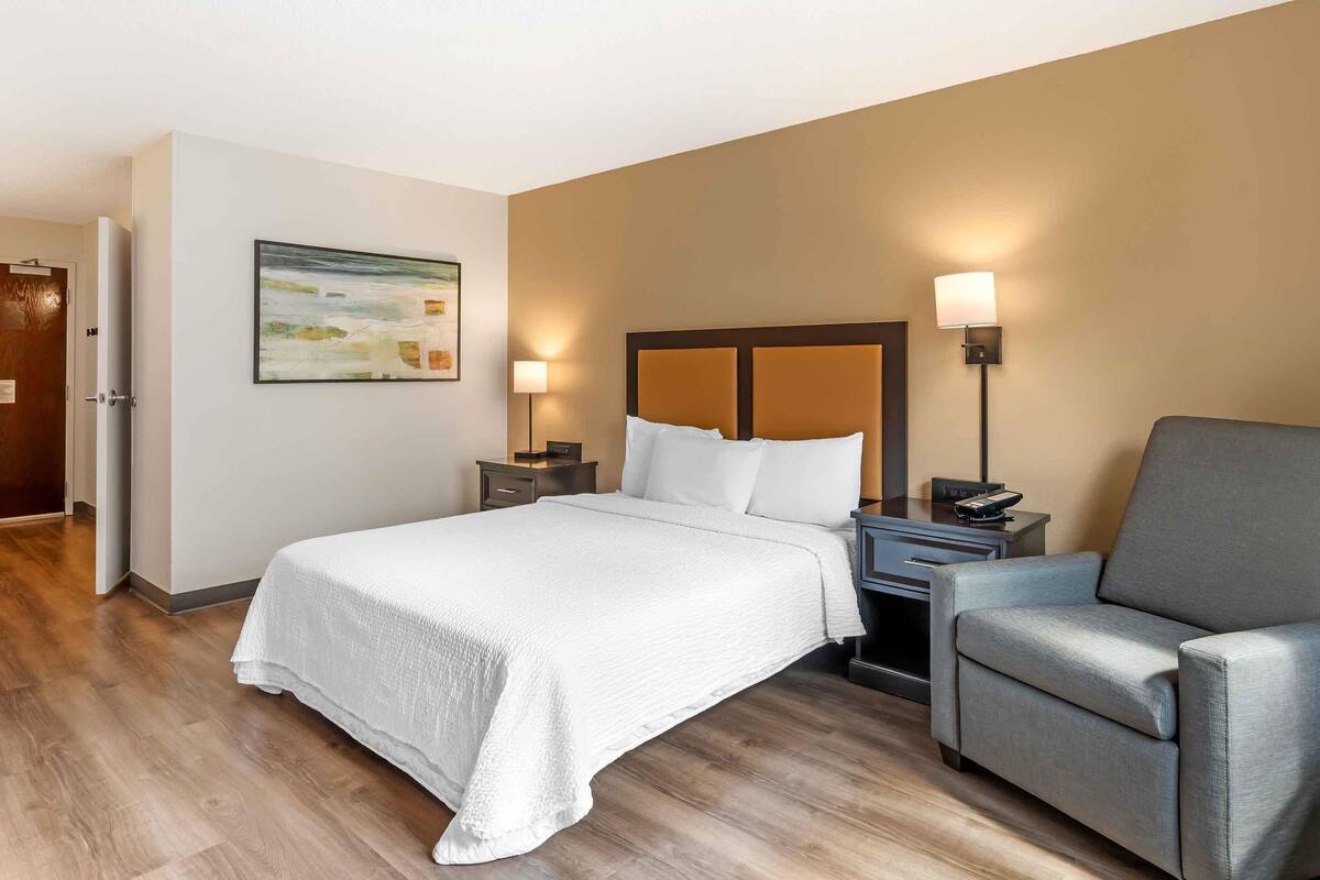 A hotel room with a bed, a chair, a nightstand, a lamp, and a painting on the wall.