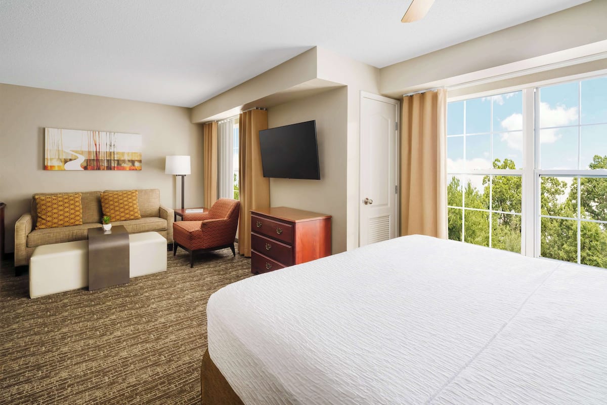 A hotel room with a large bed, a sofa, a desk, a chair, a TV, and a window with a view of trees.