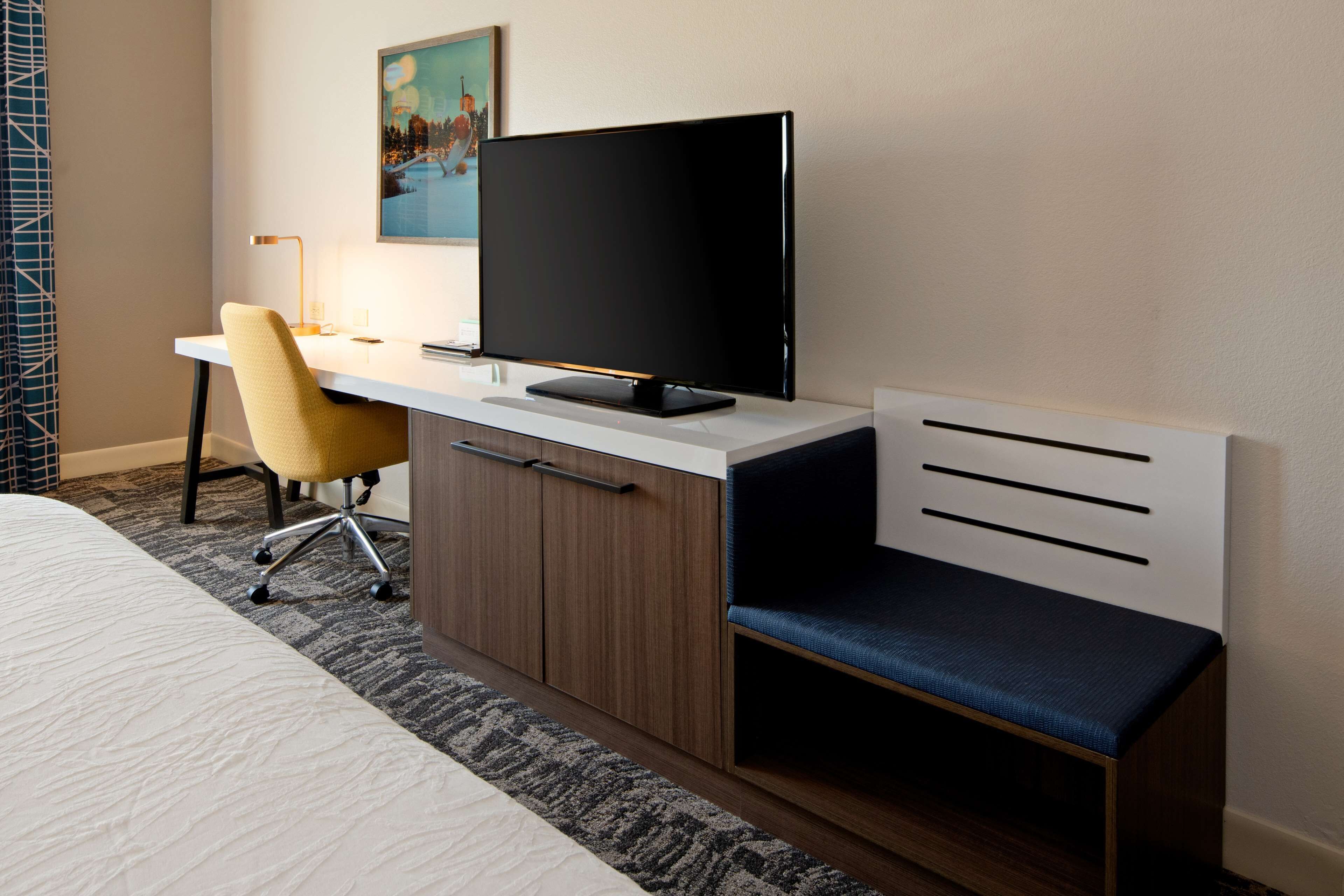 A hotel room with a bed, a desk with a computer, a chair, and a bench.