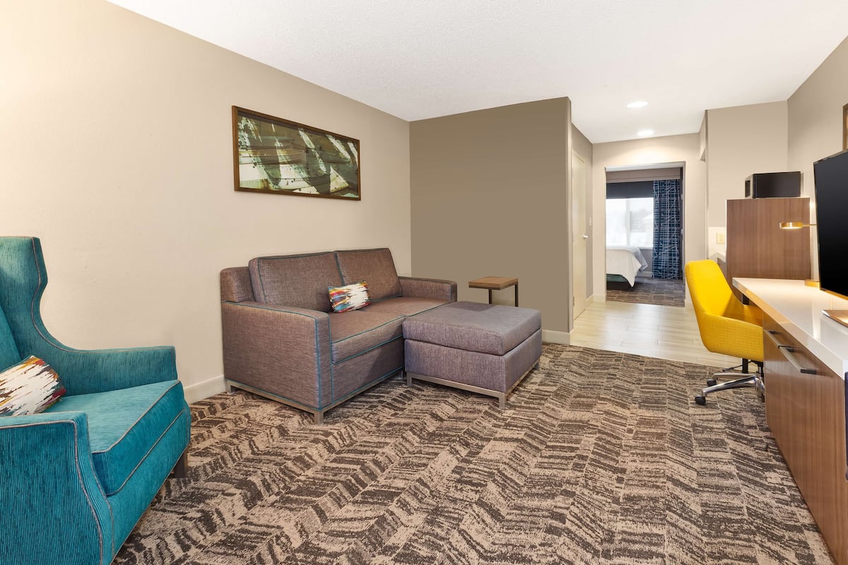 A modern hotel room with a sectional sofa, a teal armchair, a small side table, a yellow office chair, and a large window.