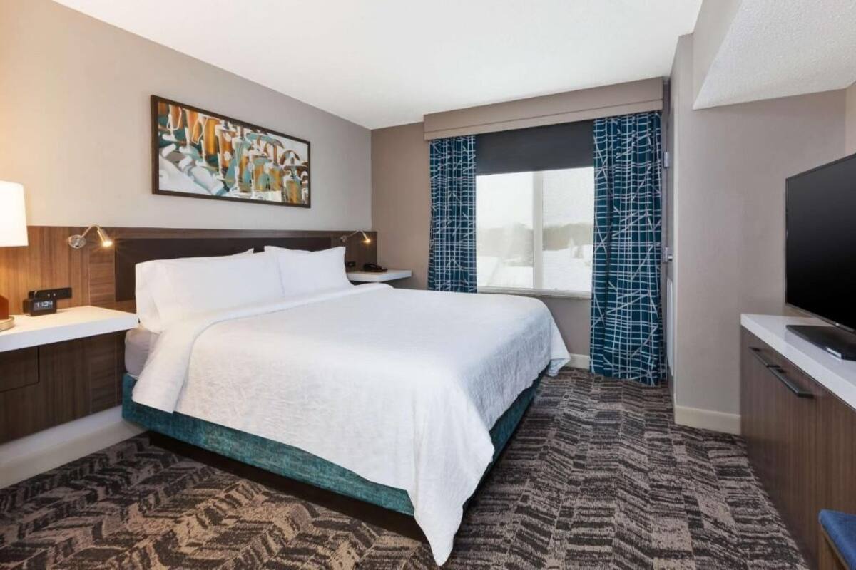 A hotel room with a bed, a TV, a painting, a window with curtains, and a patterned carpet.