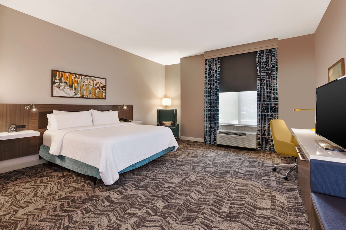A hotel room with a bed, a desk, a chair, a TV, a window, and a bathroom.