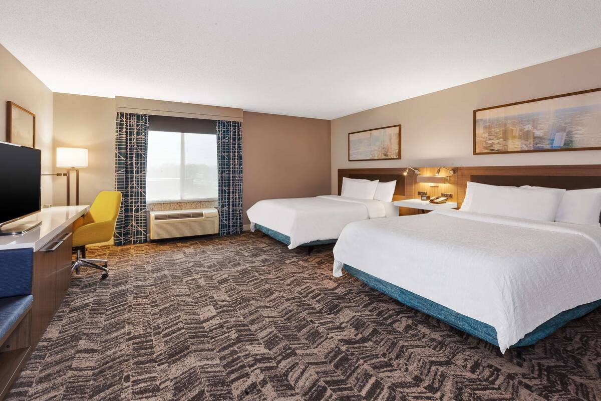 A hotel room with two beds, a desk, a chair, a TV, and a window with a view.