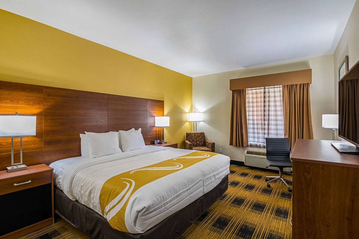 A hotel room with a large bed, a desk, a chair, a lamp, and a window with curtains.