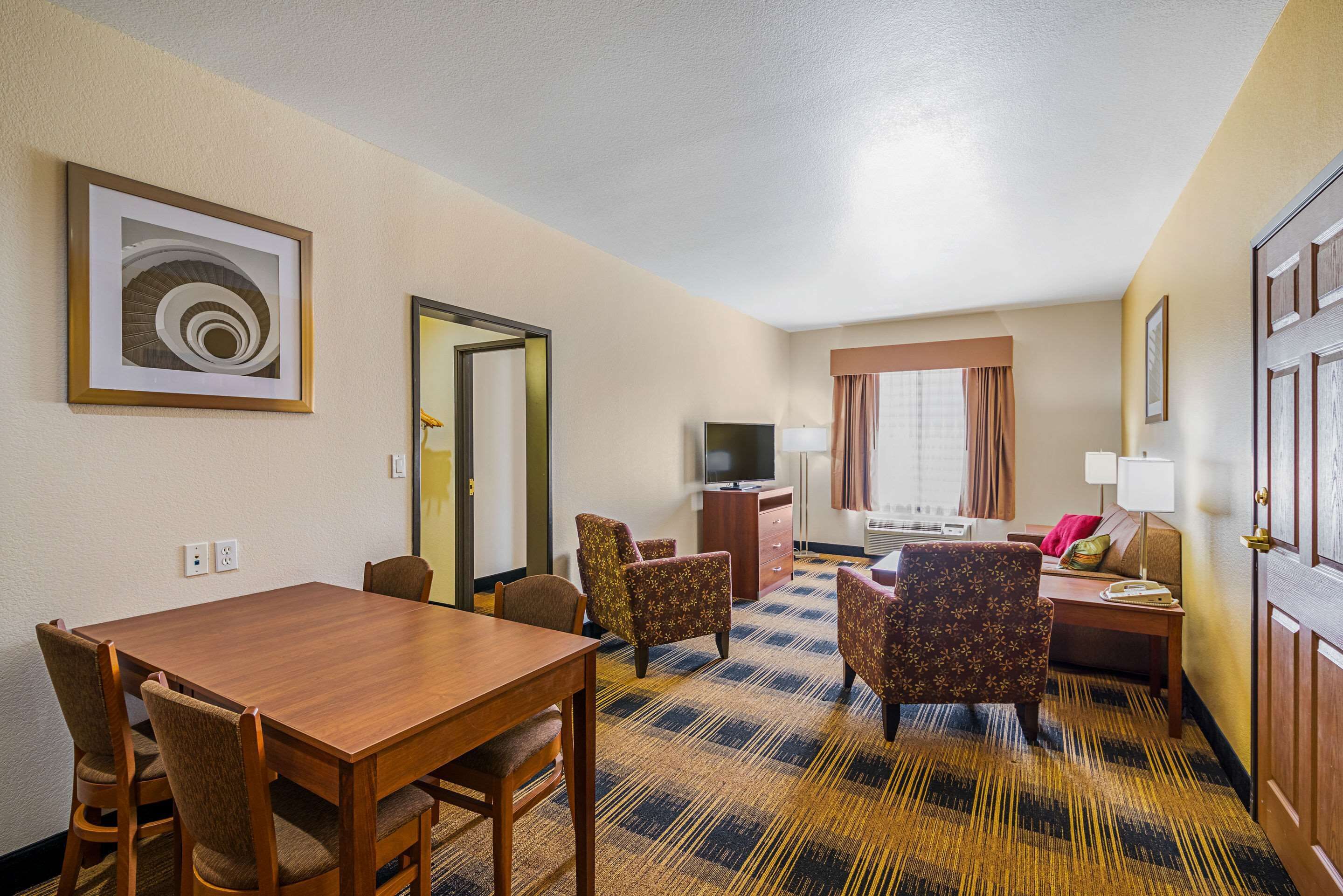 A hotel room with a wooden dining table, four chairs, a sofa, a TV, a lamp, and a patterned carpet.