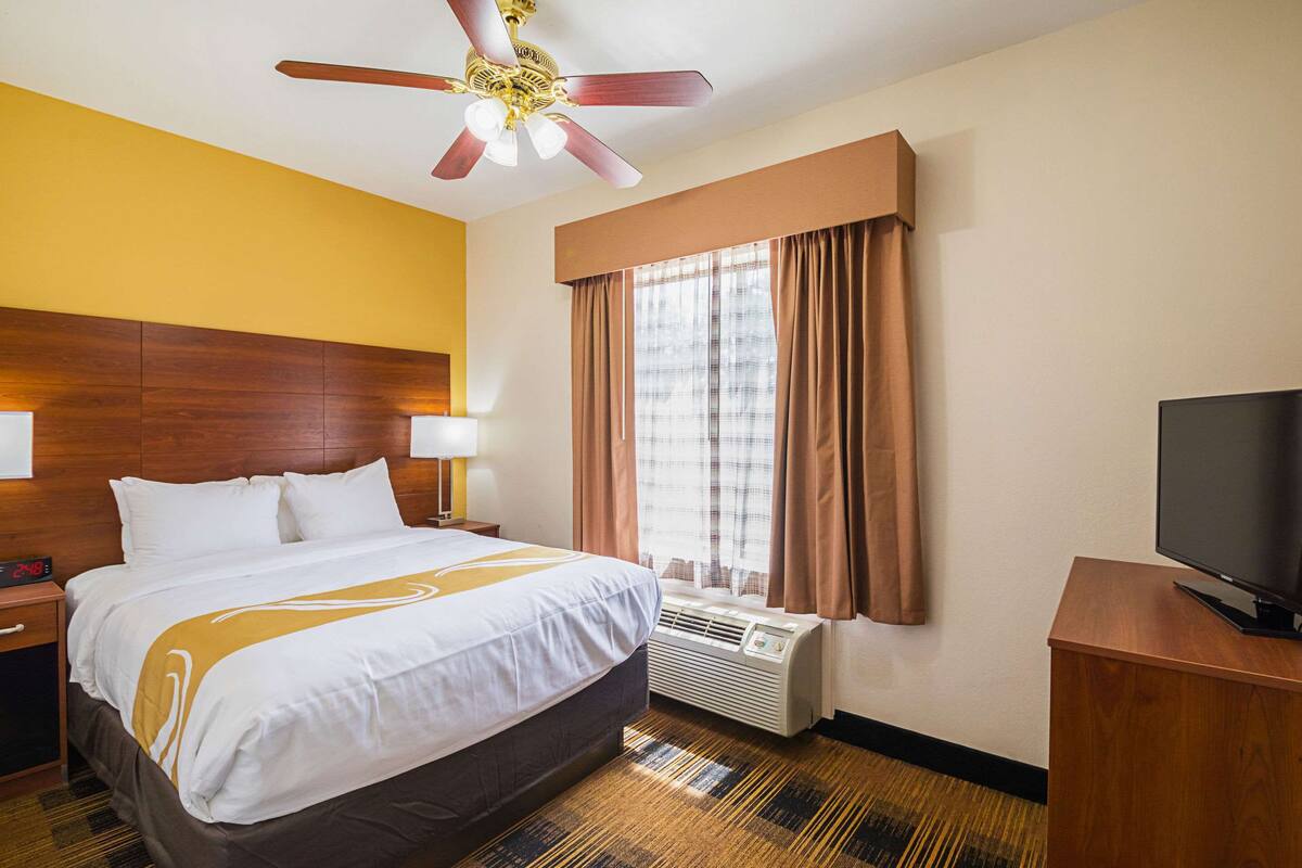 A hotel room with a bed, a ceiling fan, a TV, a window with curtains, and a wall-mounted air conditioner.