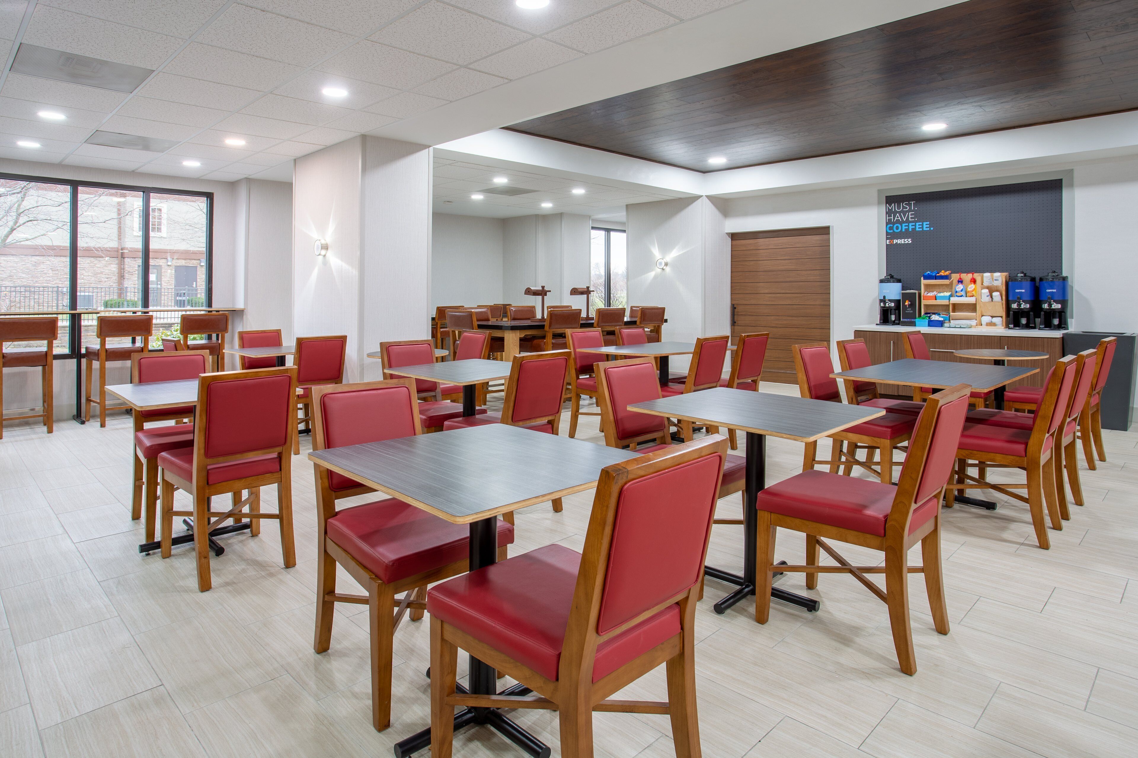 A spacious dining area with multiple tables and chairs, a coffee station, and large windows providing natural light.