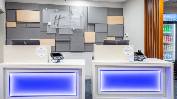 A reception area with two reception desks, each equipped with a digital kiosk and a phone. The walls are decorated with geometric patterns and a sign for 'Holiday Inn Express & Suites'. - Holiday Inn Express & Suites Fort Wayne by IHG (Fort Wayne)