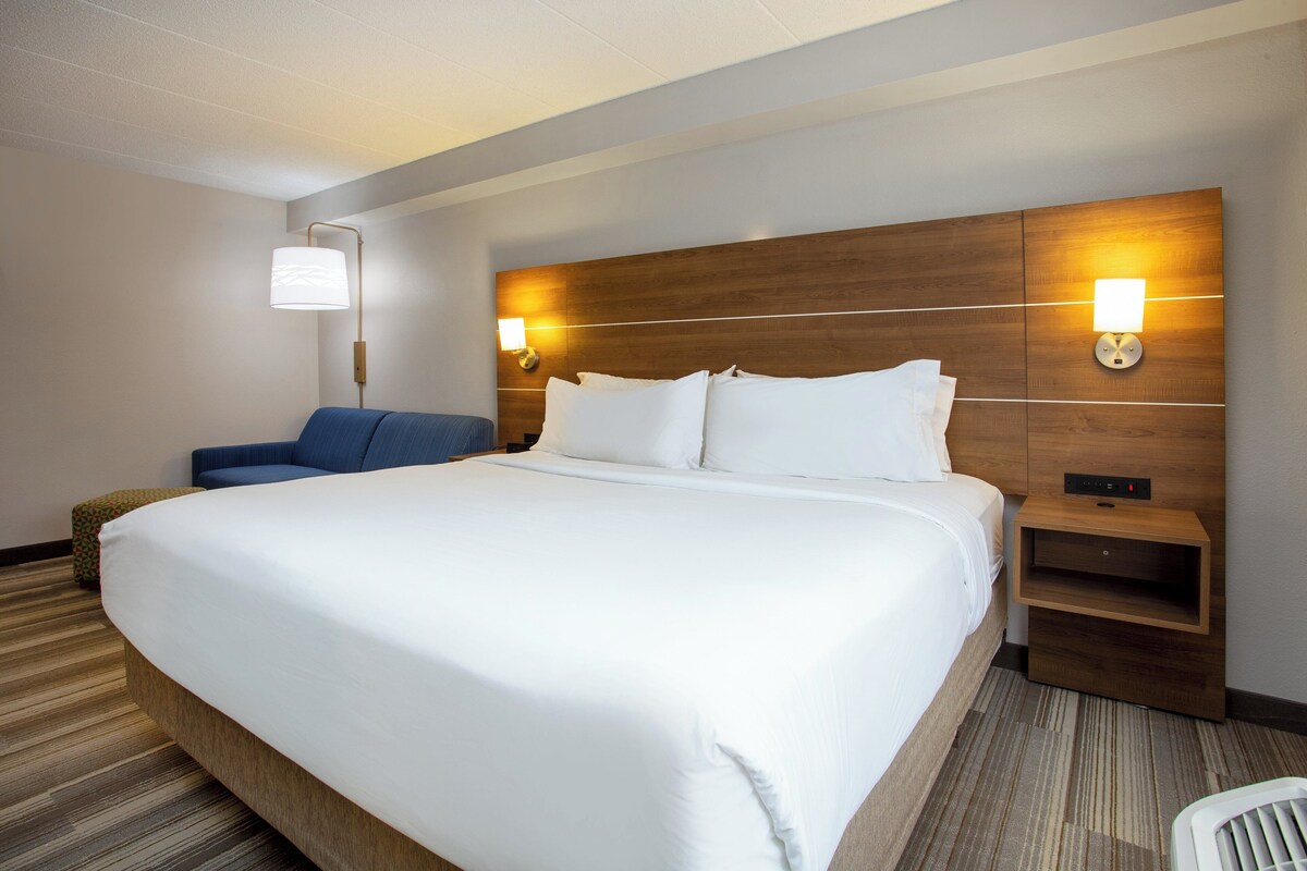 A hotel room with a large bed, a nightstand with a lamp, a blue chair, and a small ottoman.