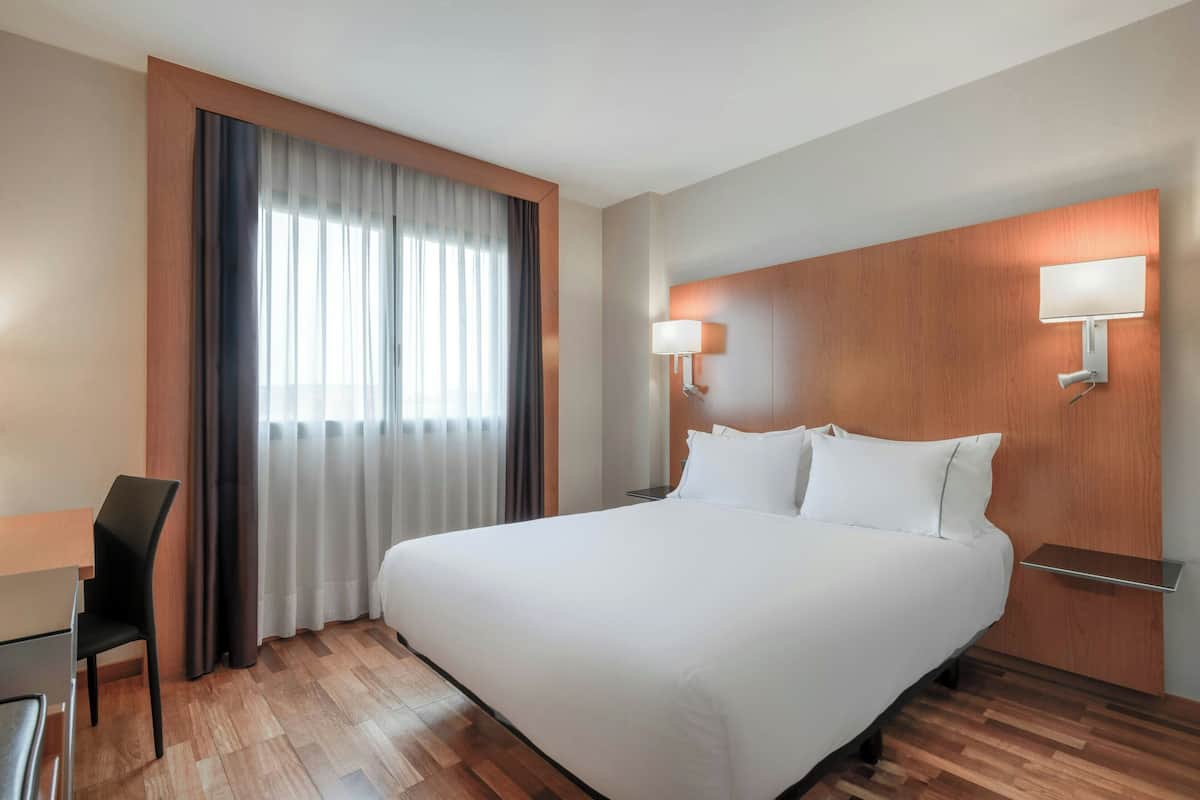Quarto de hotel com uma cama grande, uma secretária com cadeira, uma janela com cortinas e uma luminária na parede.