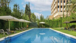 A swimming pool with lounge chairs and umbrellas, surrounded by greenery and a building in the background. - AC Hotel Sevilla Fórum by Marriott (Seville)