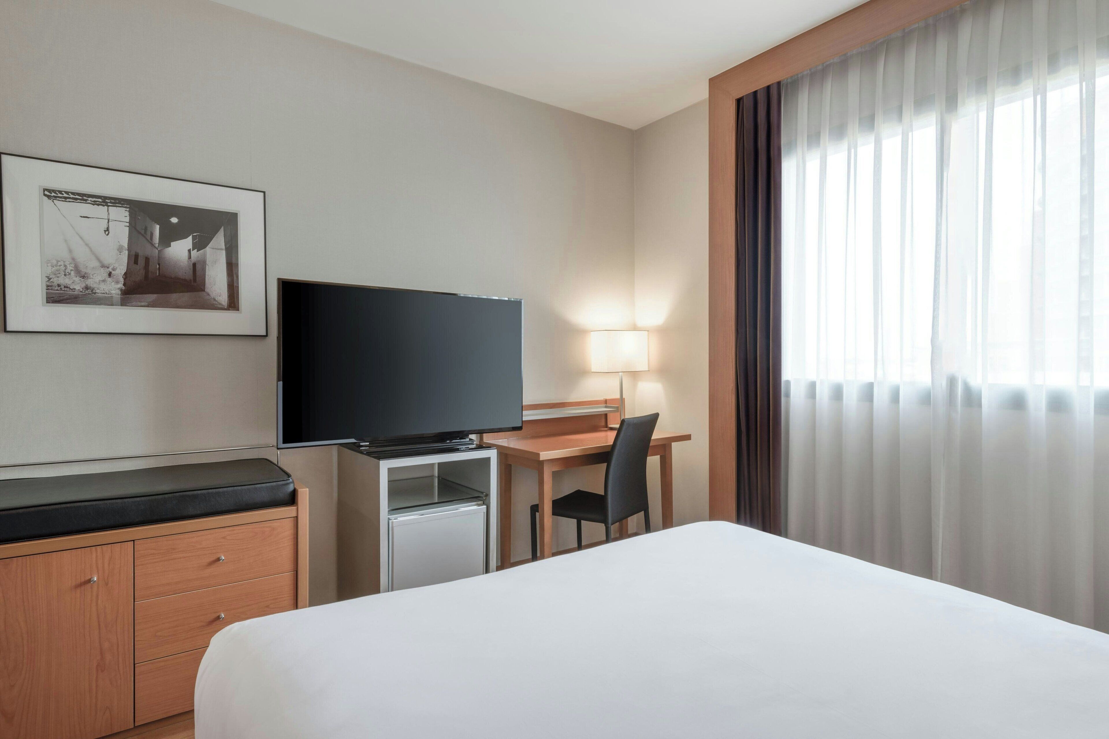 A hotel room with a bed, a desk with a chair, a television, a small refrigerator, a cabinet, and a window with curtains.