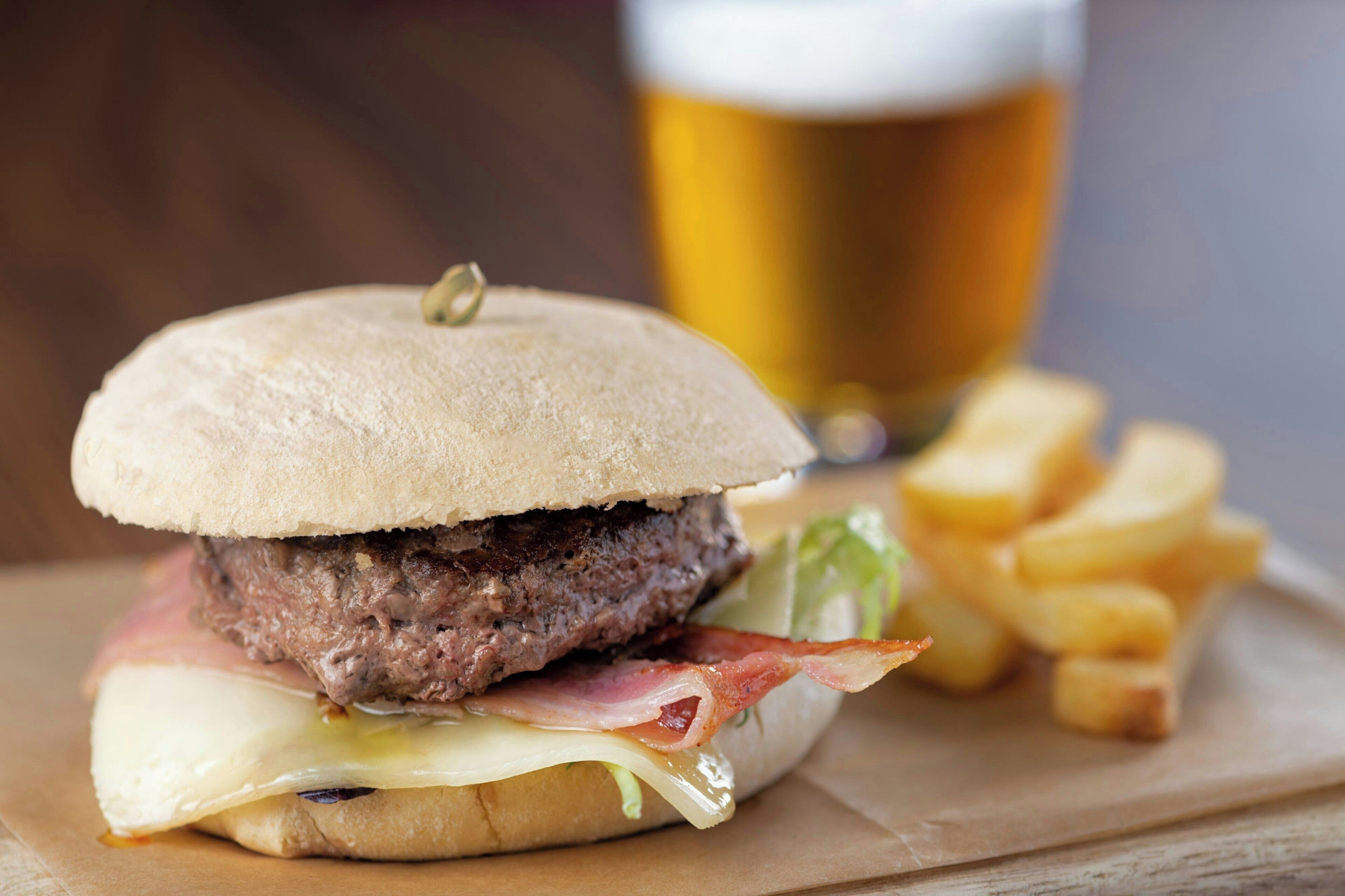 Hamburguesa con tocino, queso y lechuga en pan, acompañada de papas fritas y una jarra de cerveza.