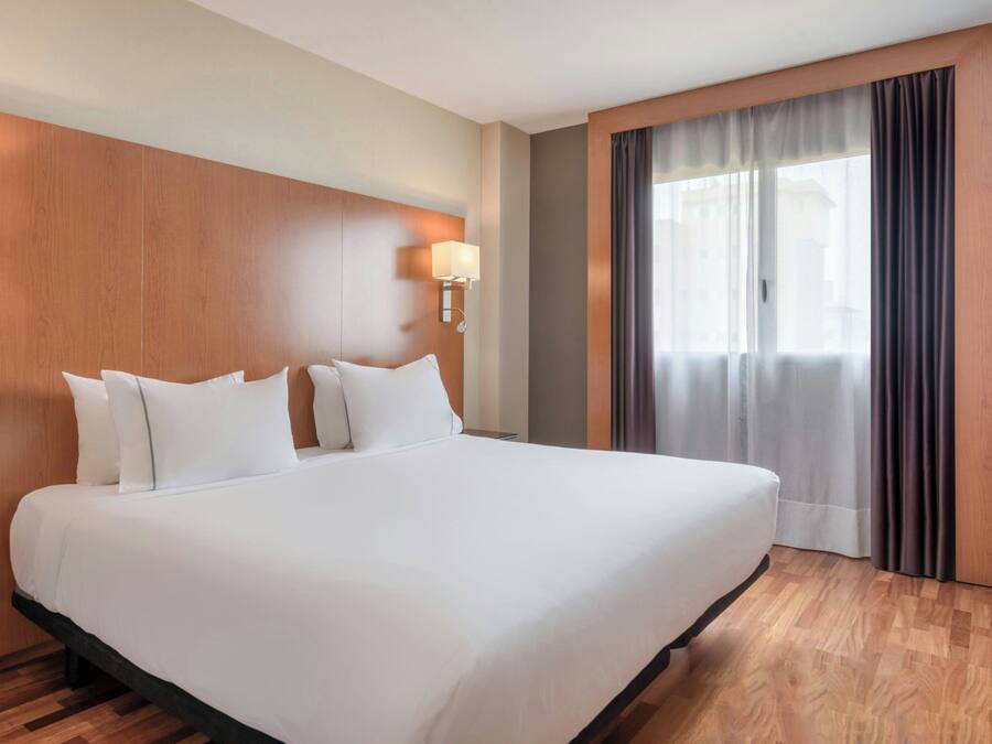 Una habitación de hotel con una cama grande, cabecera de madera, una mesita de noche, una lámpara y un ventanal con cortinas.