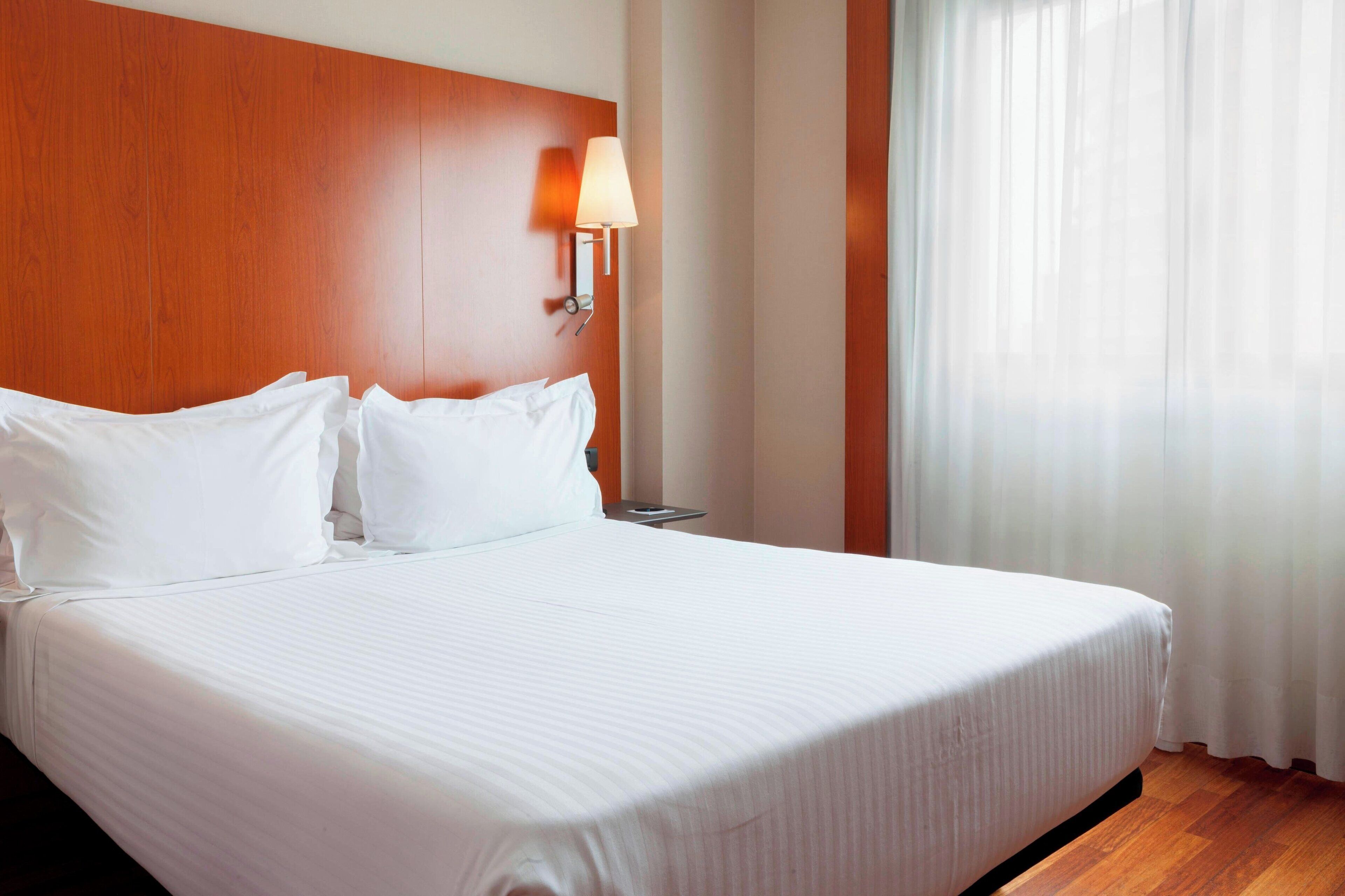 Une chambre d’hôtel avec un grand lit, une tête de lit en bois, une lampe de chevet et une fenêtre ornée de rideaux légers.