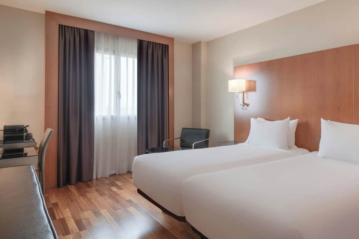 Quarto de hotel com duas camas, uma escrivaninha, uma cadeira, um abajur e uma janela com cortinas.