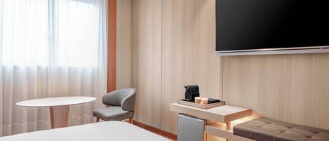 A modern hotel room with a large bed, a small round table, a grey armchair, a built-in bench, a TV mounted on the wall, and a coffee machine.