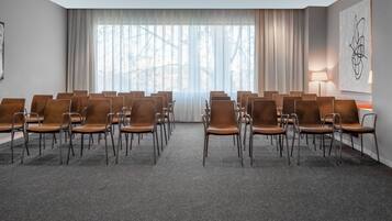 Un salón de conferencias con filas de sillas marrones, un ventanal con cortinas translúcidas, una lámpara y una obra de arte en la pared.