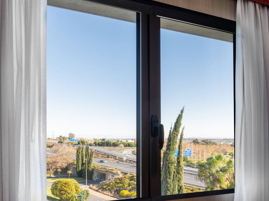 A view through a window with white curtains, revealing a road with multiple lanes, a bridge, and some greenery.