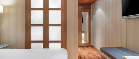 Una habitación de hotel con una cama, un banco, un televisor y una puerta de madera con paneles de vidrio esmerilado.