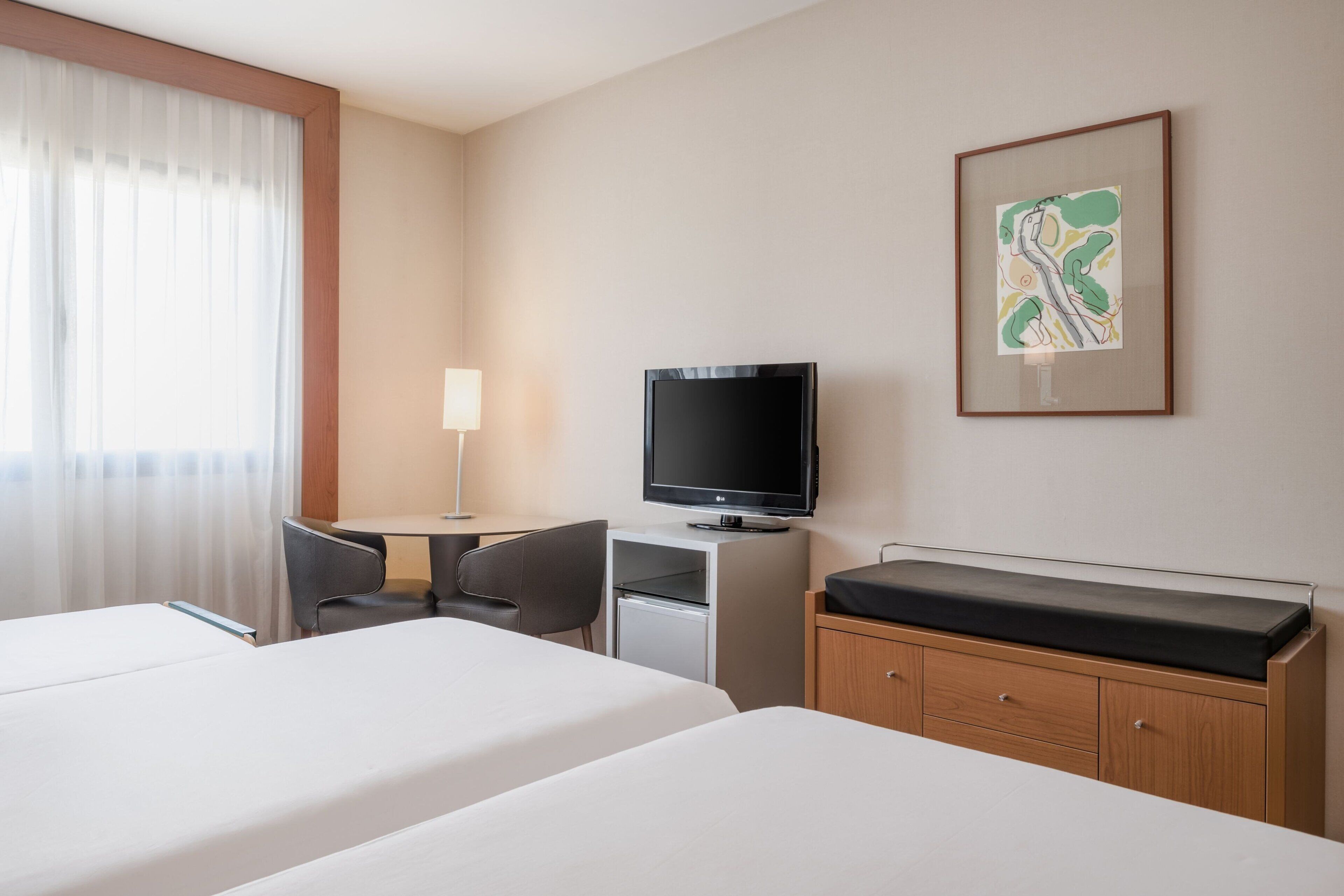 Habitación de hotel con dos camas, un escritorio con televisor, un armario pequeño y un cuadro en la pared.