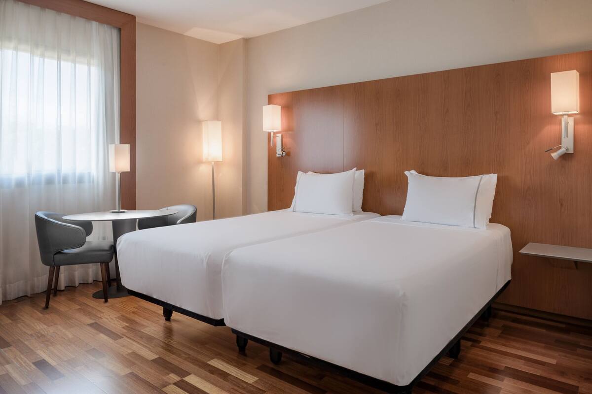 A hotel room with a double bed, two bedside tables, a small round table with two chairs, and a large window with sheer curtains.