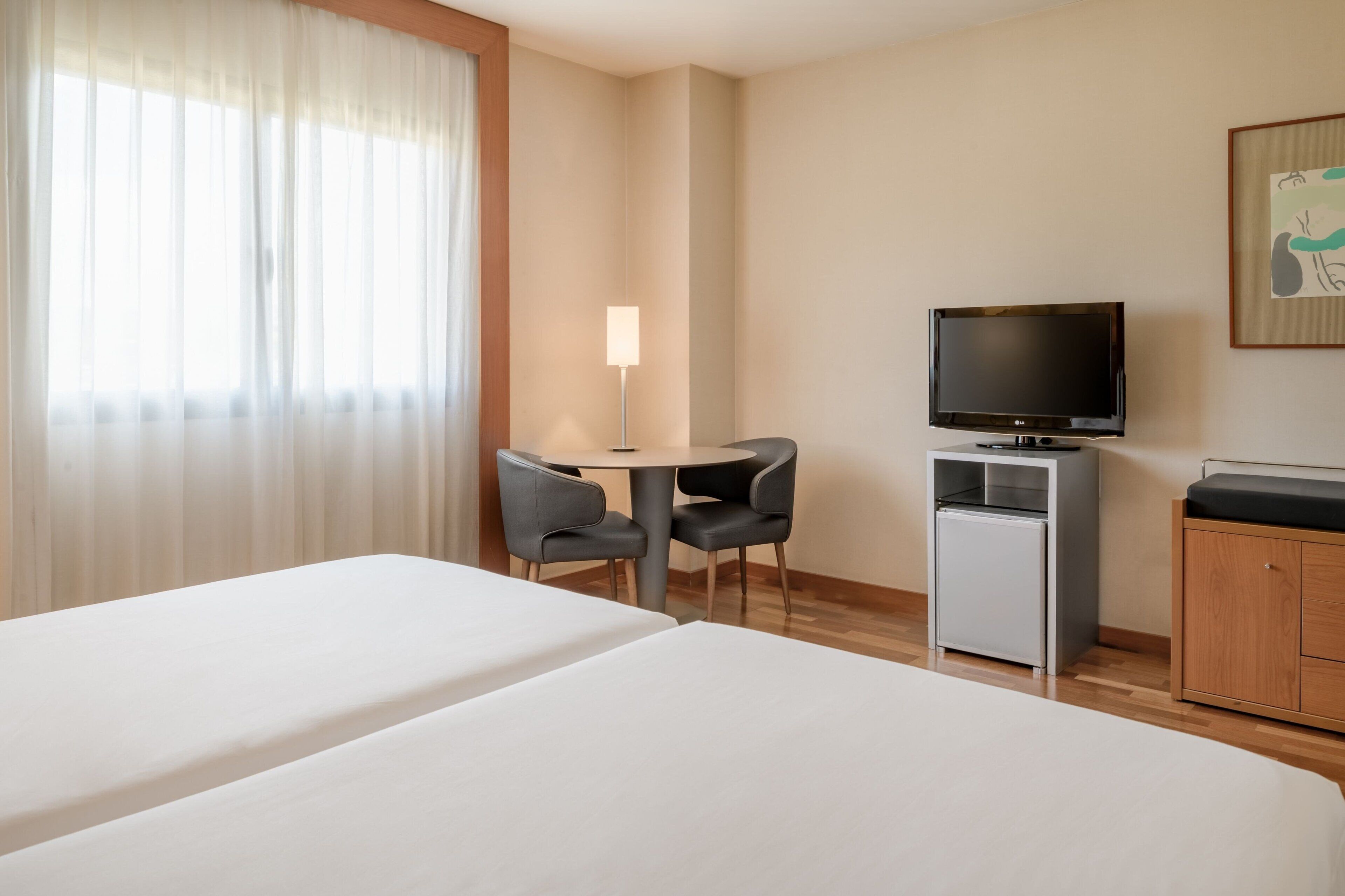 Ein Hotelzimmer mit zwei Betten, einem Schreibtisch mit Stühlen, einem Fernseher, einer Minikühlschrank und einem Holzschrank.