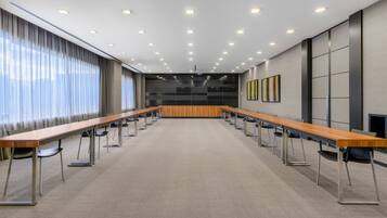 Ein Konferenzraum mit langen Tischen und Stühlen, einer hohen Decke mit eingelassener Beleuchtung und einem großen Fenster mit Vorhängen.
