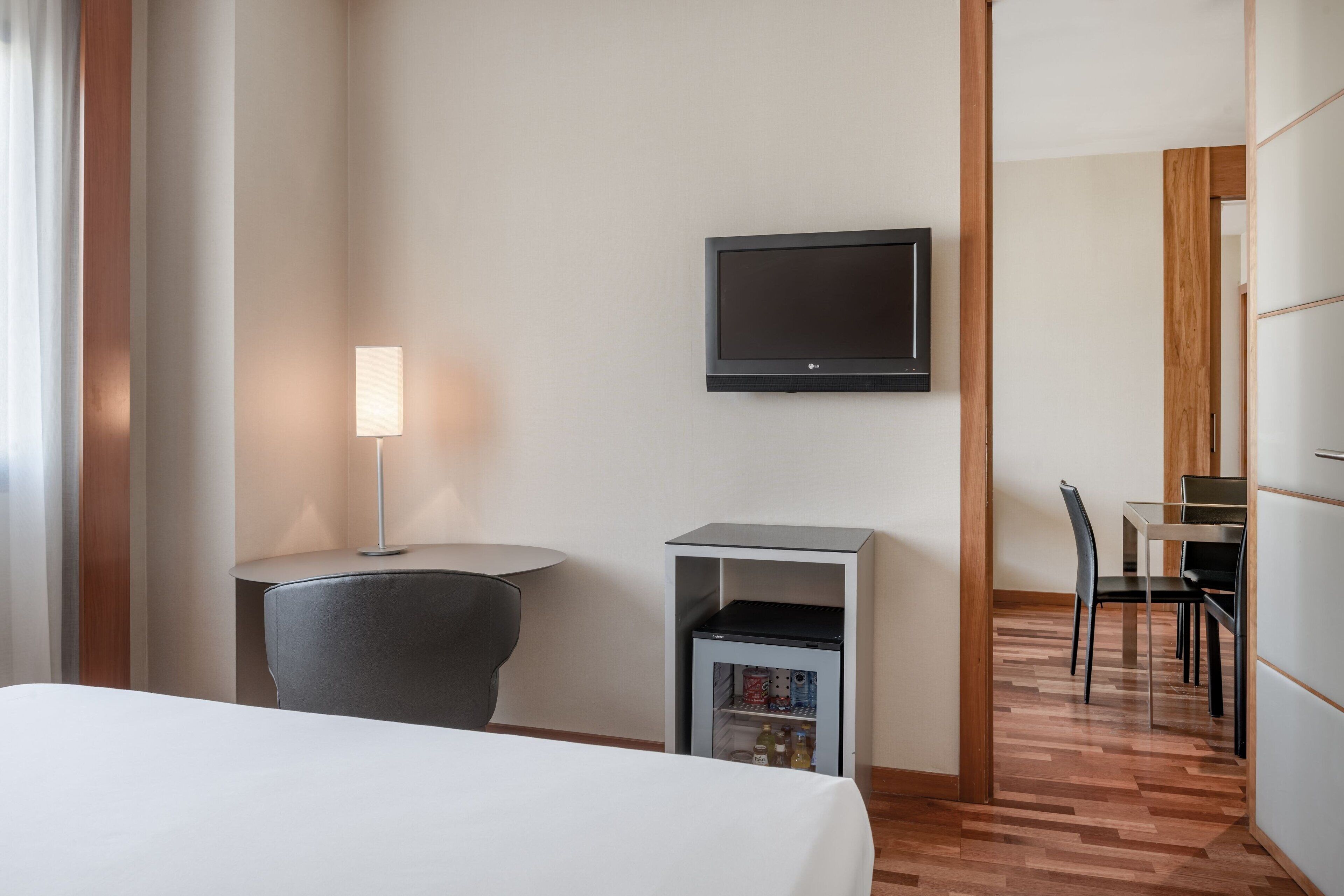 A hotel room with a bed, a desk with a chair, a television mounted on the wall, and a small refrigerator.