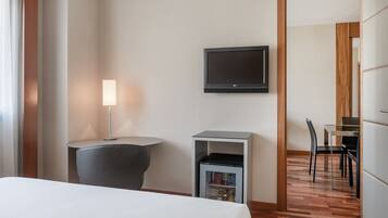 A hotel room with a bed, a desk with a chair, a television mounted on the wall, and a small refrigerator.
