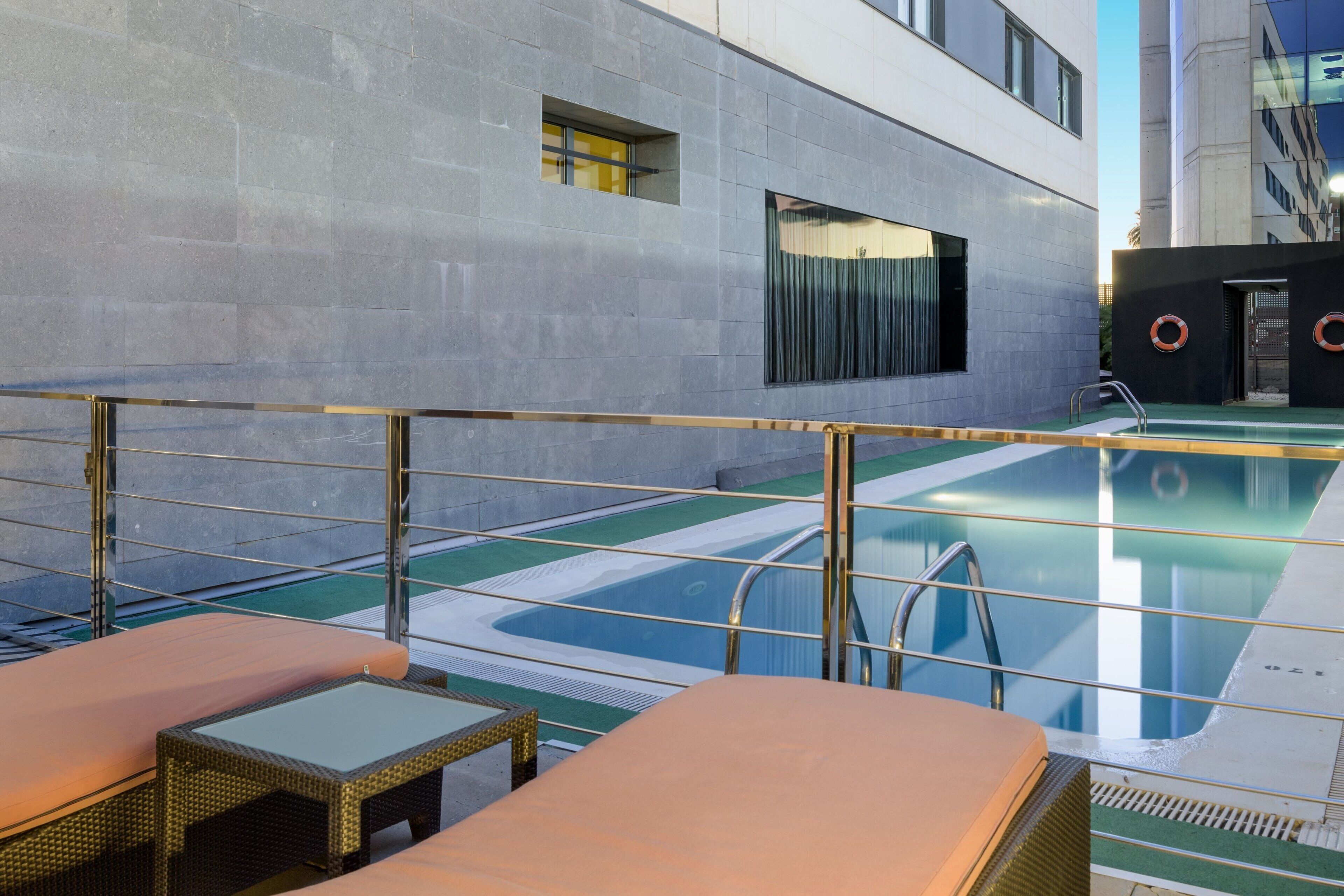 Een dakzwembad met ligbedden, een glazen balustrade en uitzicht op een modern gebouw.