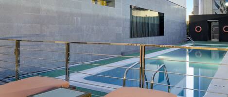 Een dakzwembad met ligbedden, een glazen balustrade en uitzicht op een modern gebouw.