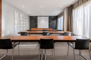 Un salón de conferencias con una larga mesa, sillas negras, una pantalla de proyector y un amplio ventanal con cortinas.
