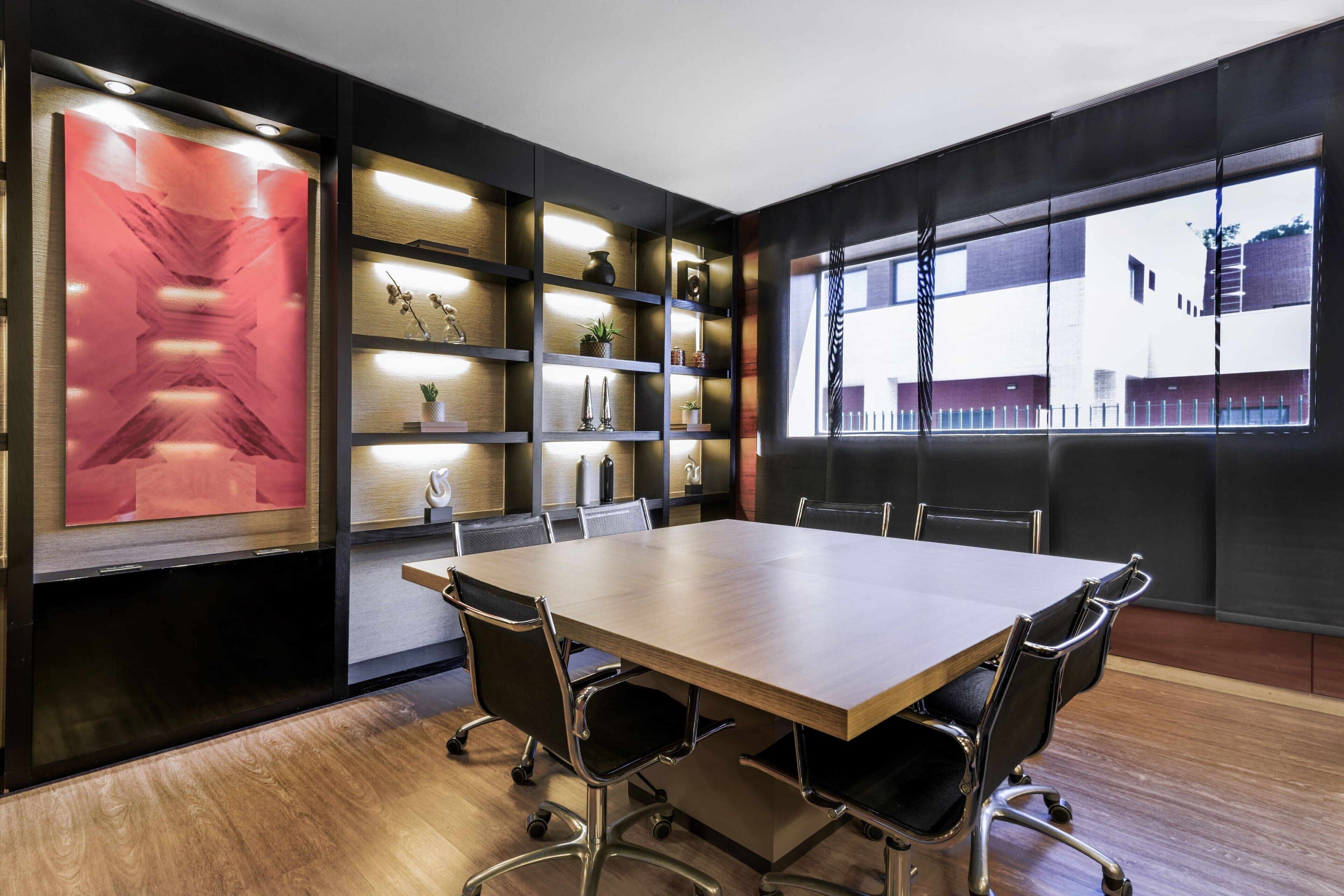 A conference room with a large table, chairs, shelves with decorative items, and a view through a window.