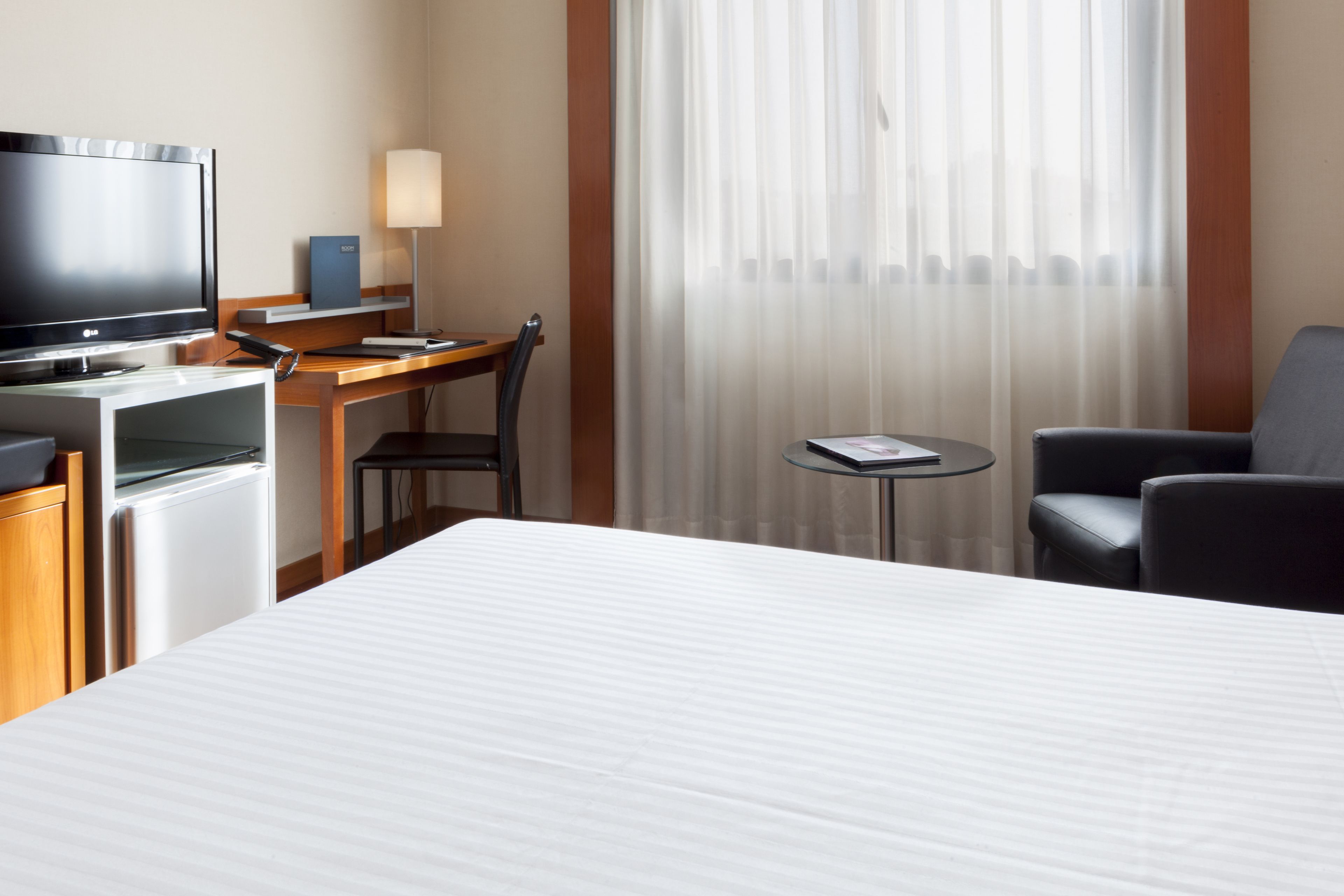 Habitación de hotel con cama, escritorio con computadora, televisor, lámpara y ventana con cortinas traslúcidas.