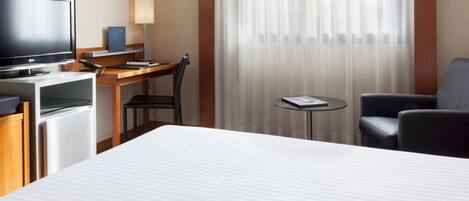 A hotel room with a bed, a desk with a computer, a television, a lamp, and a window with sheer curtains.