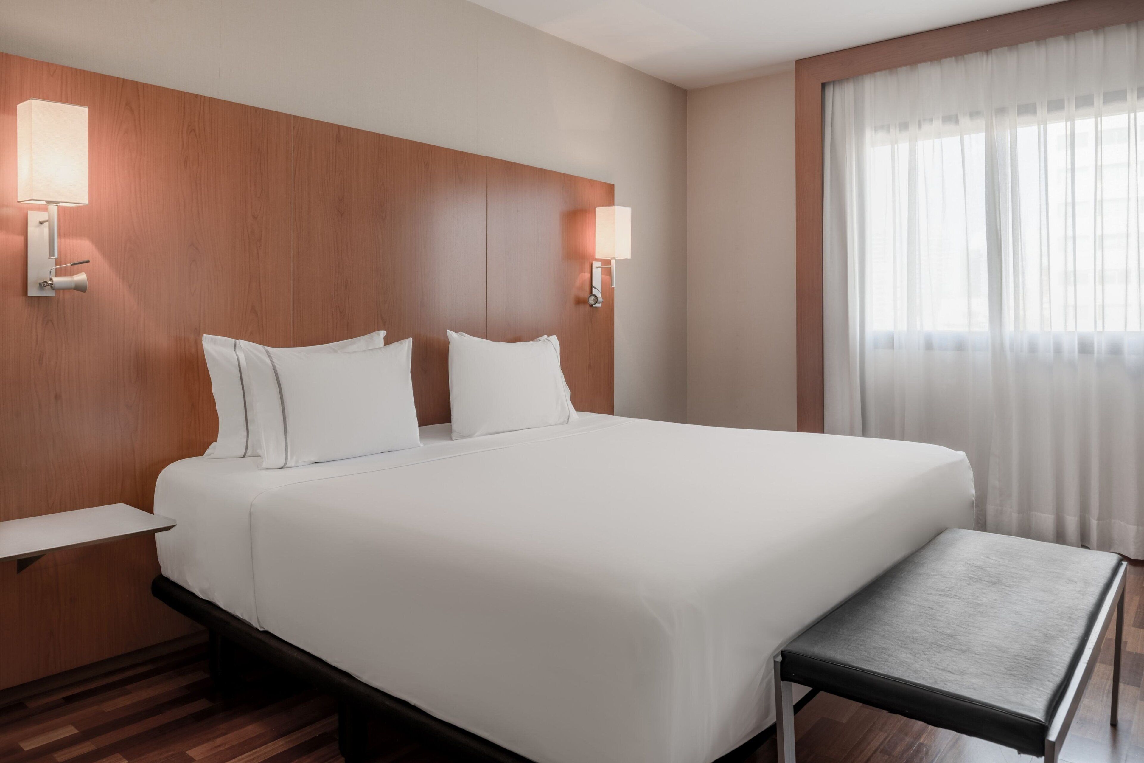 Habitación de hotel con una cama grande, dos mesitas de noche, un banco al pie de la cama y dos lámparas de pared.