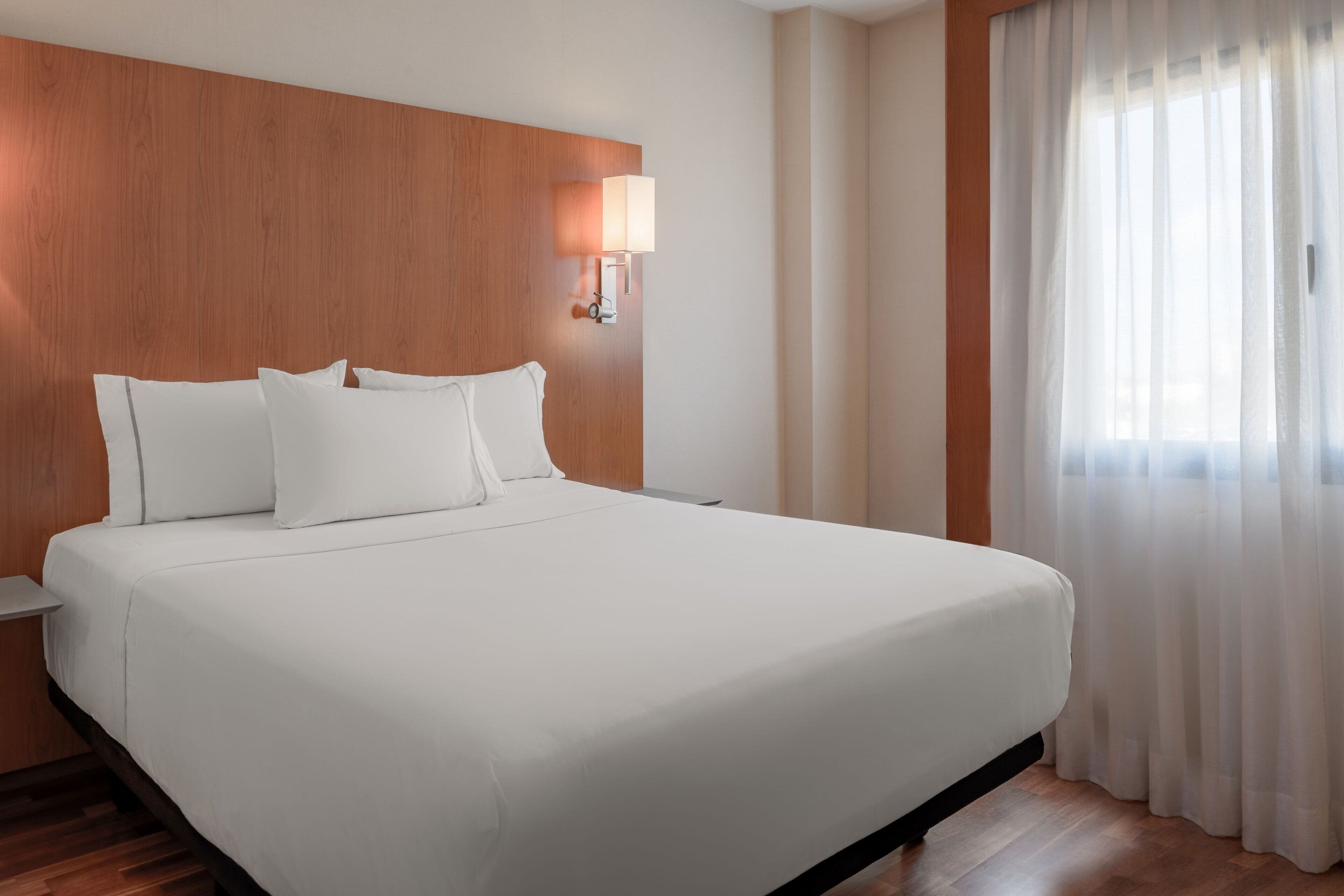 Habitación de hotel con una cama grande, cabecera de madera, ropa de cama blanca, una mesita de noche, una lámpara de pared y cortinas de gasa.