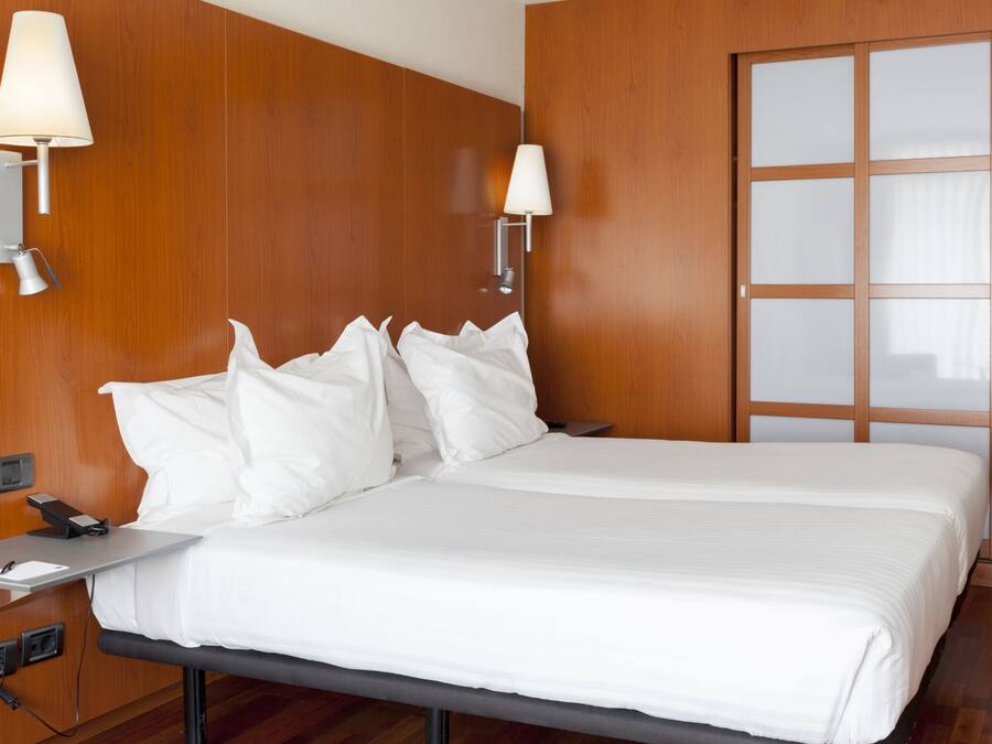 Habitación de hotel con pared de madera, cama con ropa blanca, dos lámparas de noche y una ventana con paneles de vidrio esmerilado.