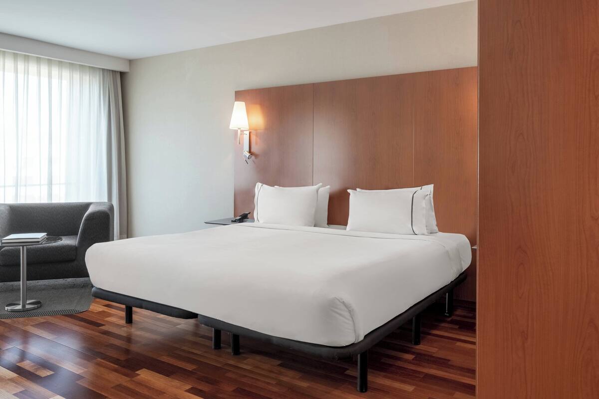 A hotel room with a large bed, a bedside table, a lamp, a sofa, and a window with curtains.