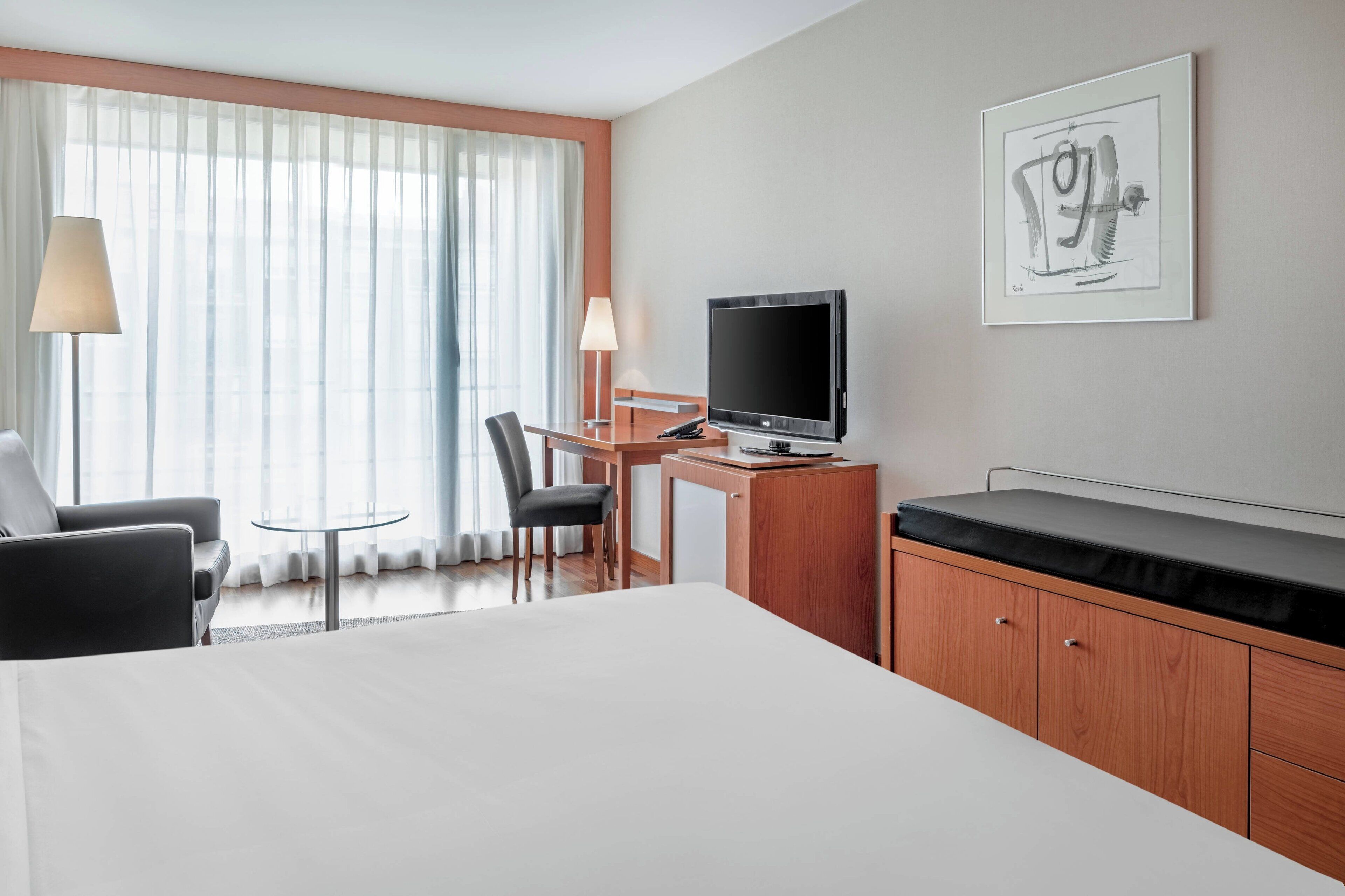 Habitación de hotel con una cama grande, un escritorio con computadora, una silla, televisión y ventana con cortinas traslúcidas.