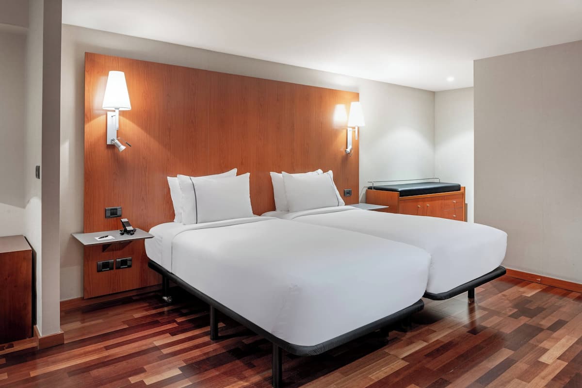 A hotel room with two beds, wooden headboard, bedside lamps, and a small wooden nightstand.