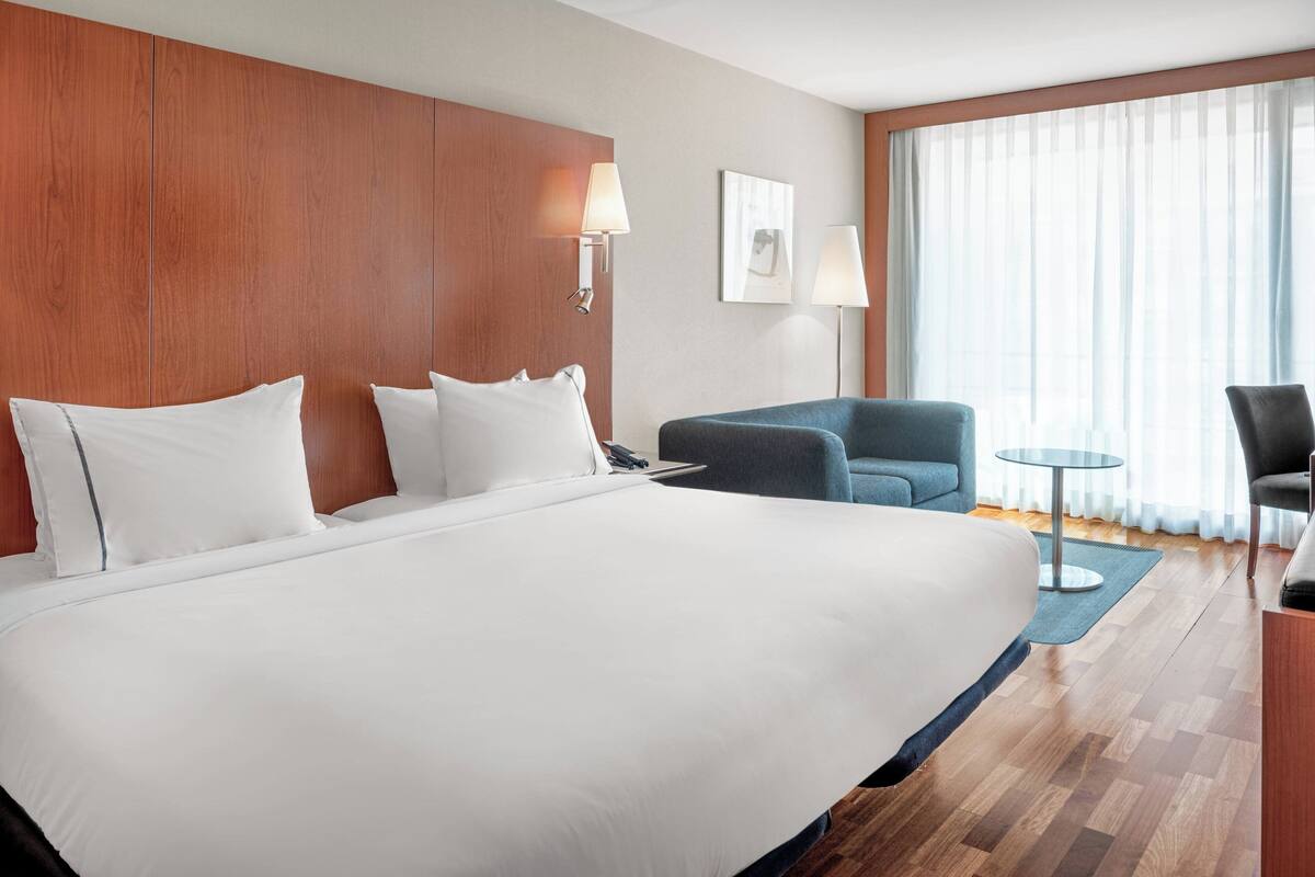 A hotel room with a large bed, a desk with a chair, a small table, and a large window with curtains.