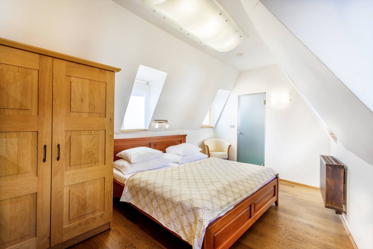 A bedroom with a wooden wardrobe, a bed with a patterned bedspread, a chair, and a radiator.