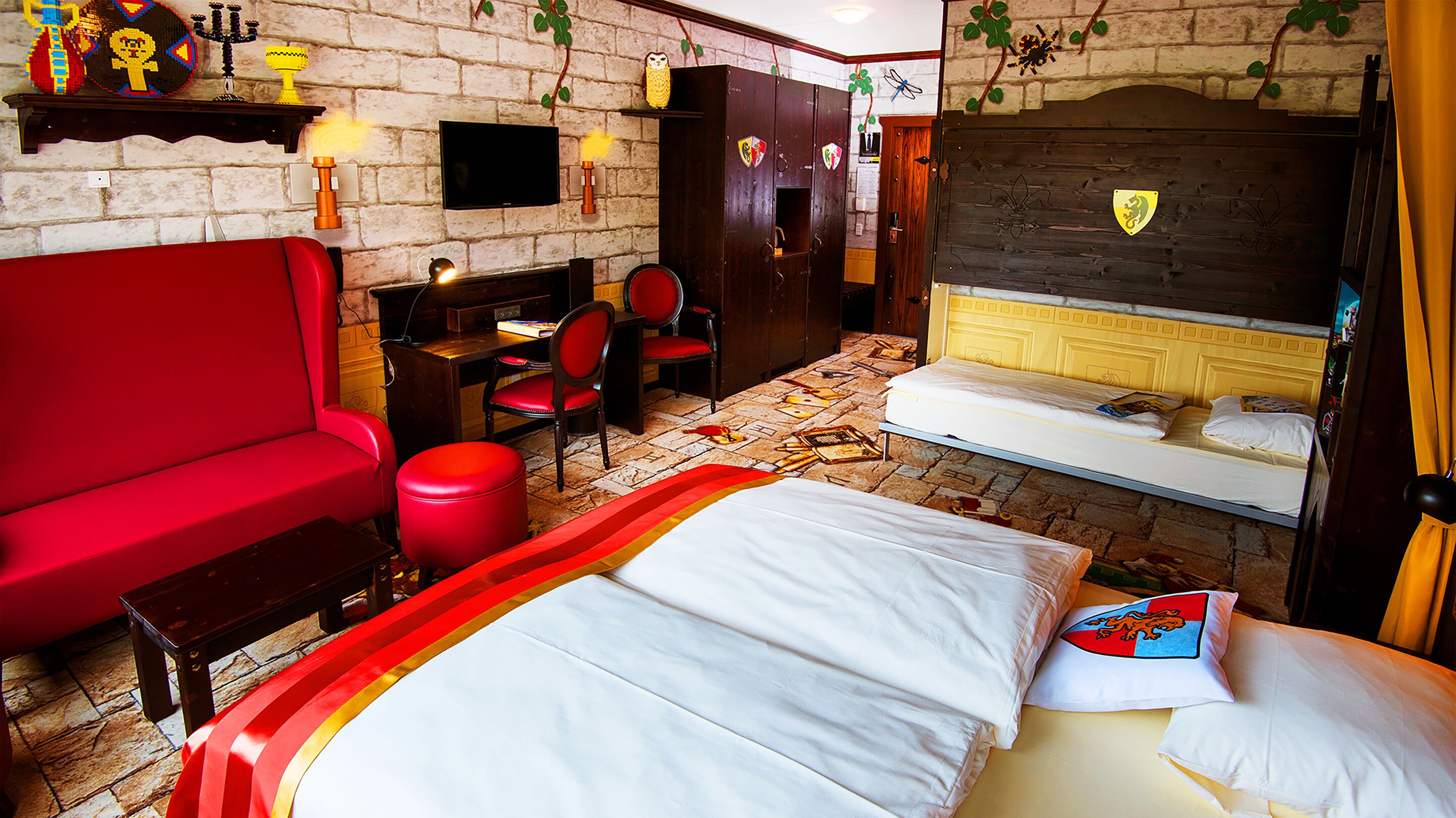A hotel room with a red sofa, a bed with white bedding, a desk with a chair, and a television on the wall.