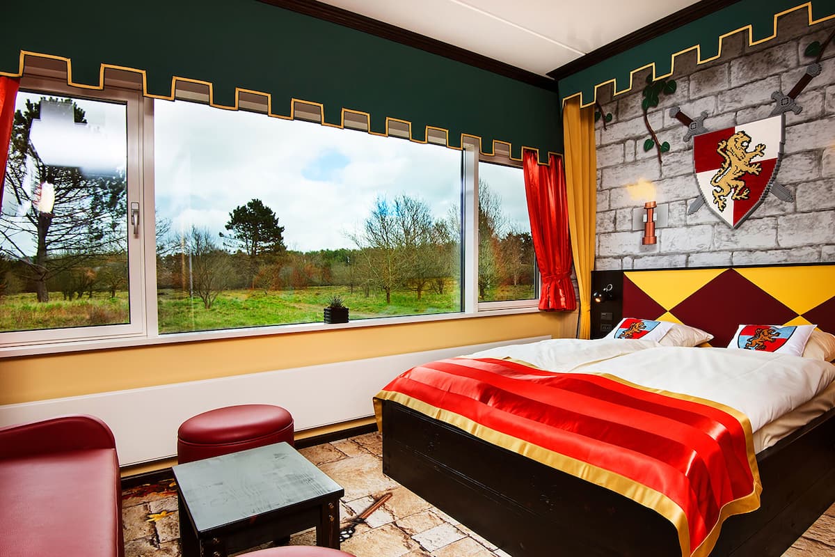 A bedroom with a large window, a bed with a red and gold bedspread, and a stone wall with a coat of arms.