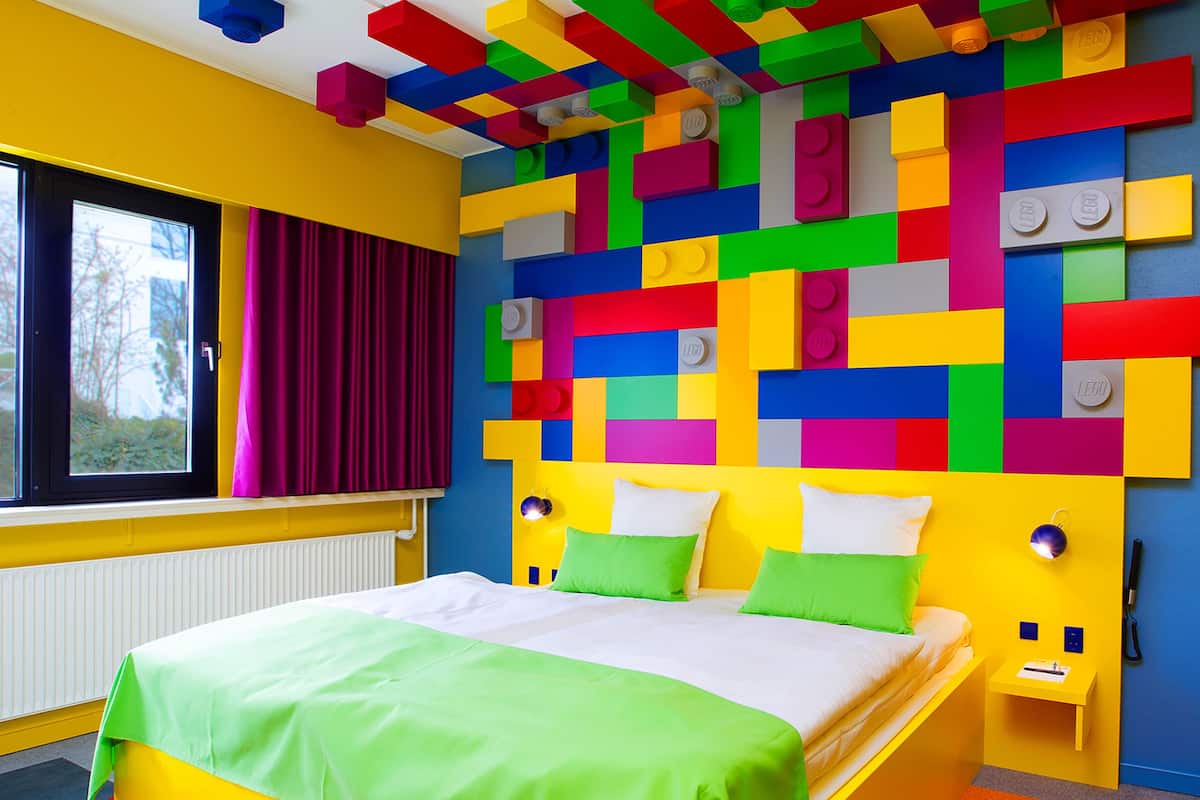 A brightly colored room with a bed, a window with curtains, and a wall decorated with LEGO bricks.