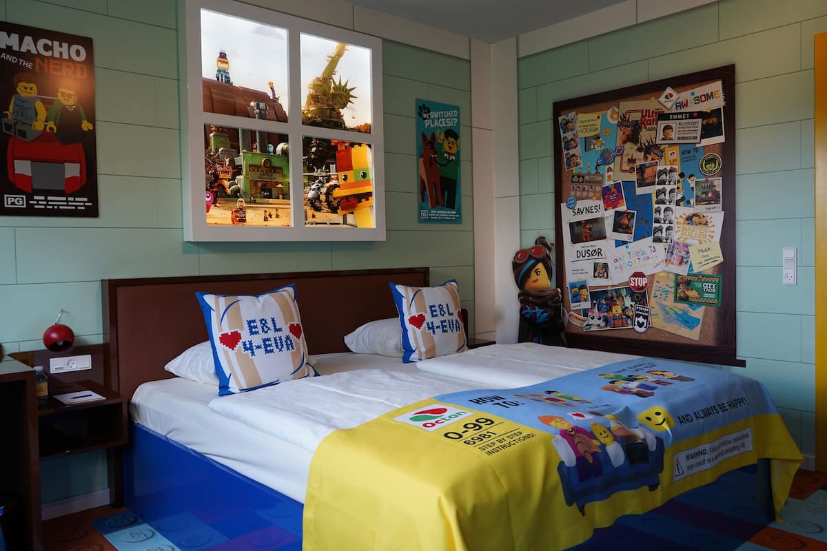 A hotel room with a bed, a nightstand, a window with a view of a building, and a bulletin board on the wall.